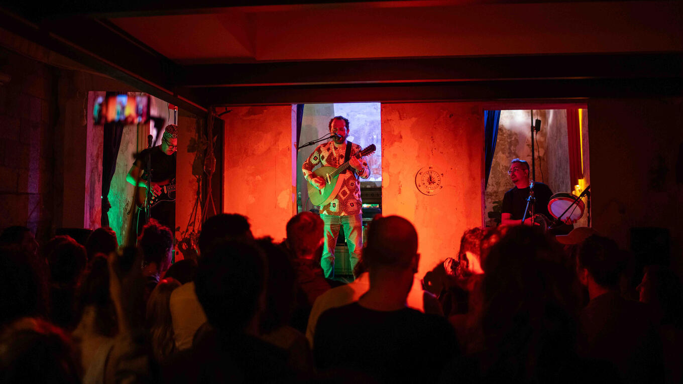 Café Noailles performs a vibrant North African-inspired set in a warm, red-lit room. The crowd is tightly packed, swaying and dancing, with one musician center stage on mandolin, flanked by electric guitar and percussion.