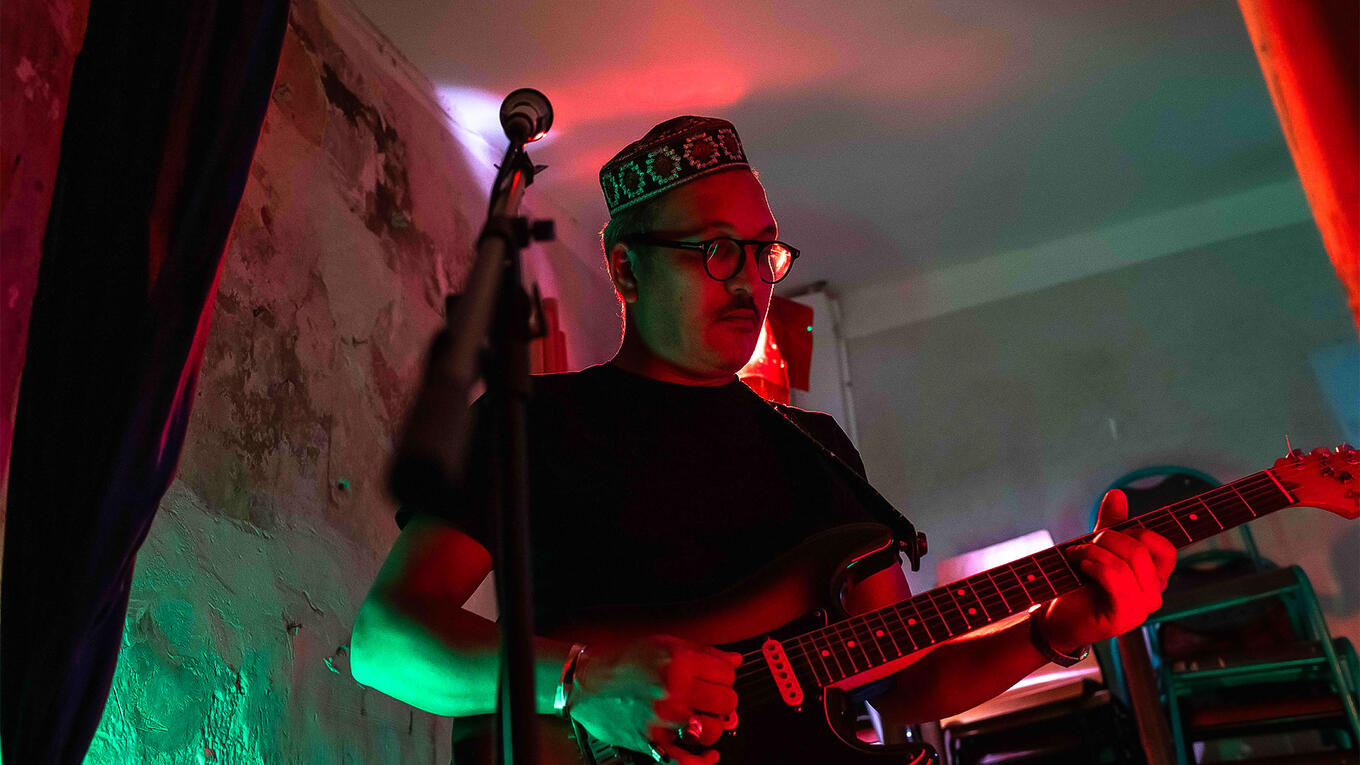 Djamel Reffes plays electric guitar under red and green lights, wearing a patterned kufi hat and glasses, focused mid-performance.