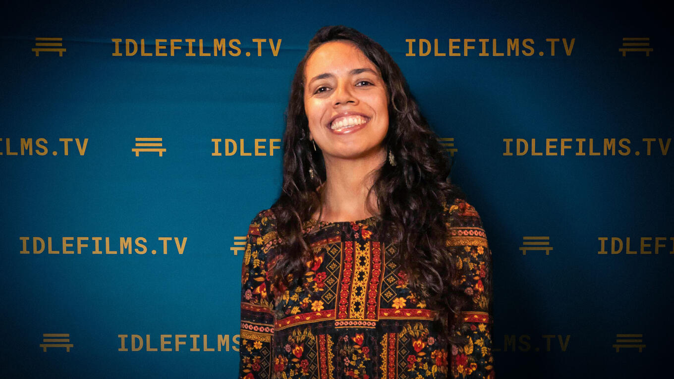 Portrait of a smiling Brazilian actress in a patterned dress, standing in front of the Idle Films TV backdrop.