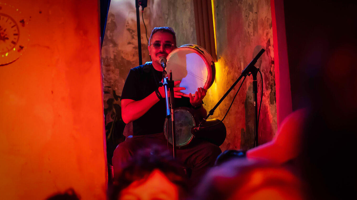 Musician Mehdi Laifaoui plays a North African frame drum on stage, lit by warm red and yellow light, seated with a mic in front.