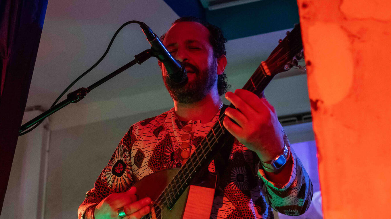 Kader Denedia sings into a microphone while playing a stringed instrument, lit by colorful stage lighting during a live performance.