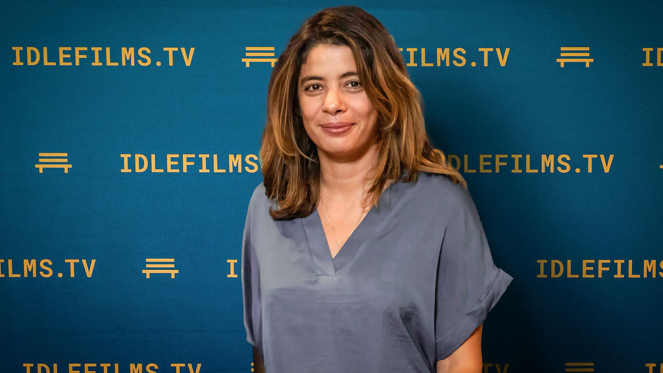 Portrait of actress Naziha Hormani standing in front of the Idle Films TV backdrop, smiling at the camera.