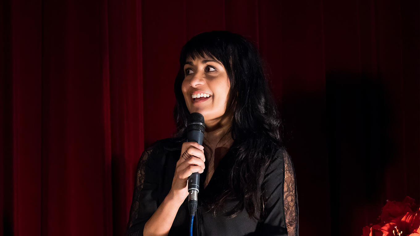 Mala Ghedia moderating the Hamburg premiere Q&A, holding a microphone in front of a red curtain.