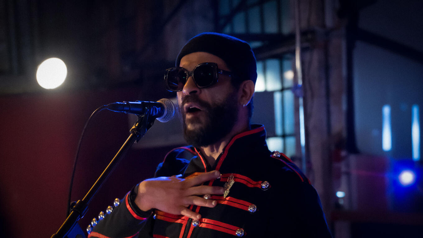 Mustapha Lakhdari at the microphone, wearing sunglasses and a red-trimmed black jacket, reciting poetry at the Idle Films event.