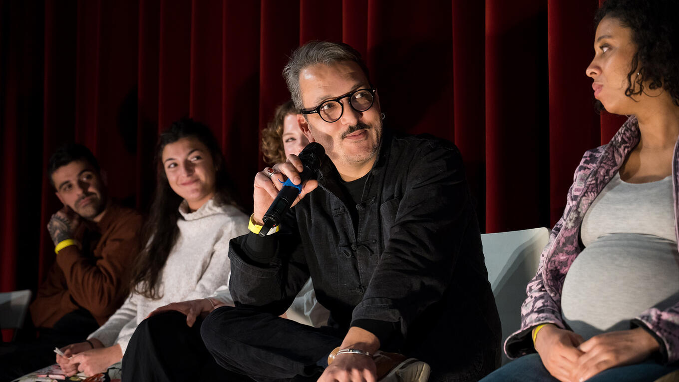 Djamel Reffes, actor and musician featured in Sunny Side Up, answers a question during the Idle Films premiere Q&A, seated among cast and crew in front of a red curtain.